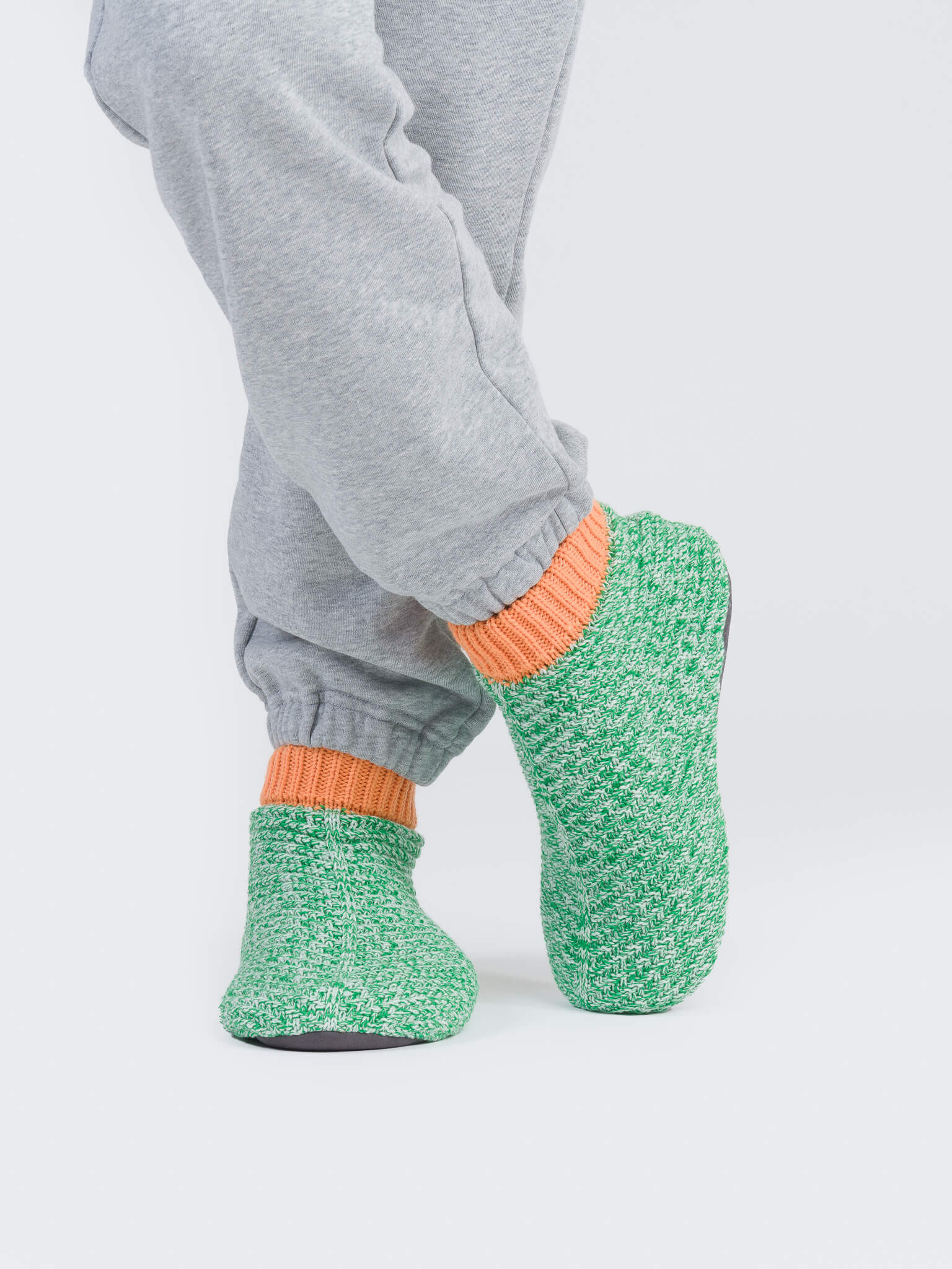 A person stands against a plain white background, wearing gray sweatpants and orange socks paired with cozy green knit slippers.