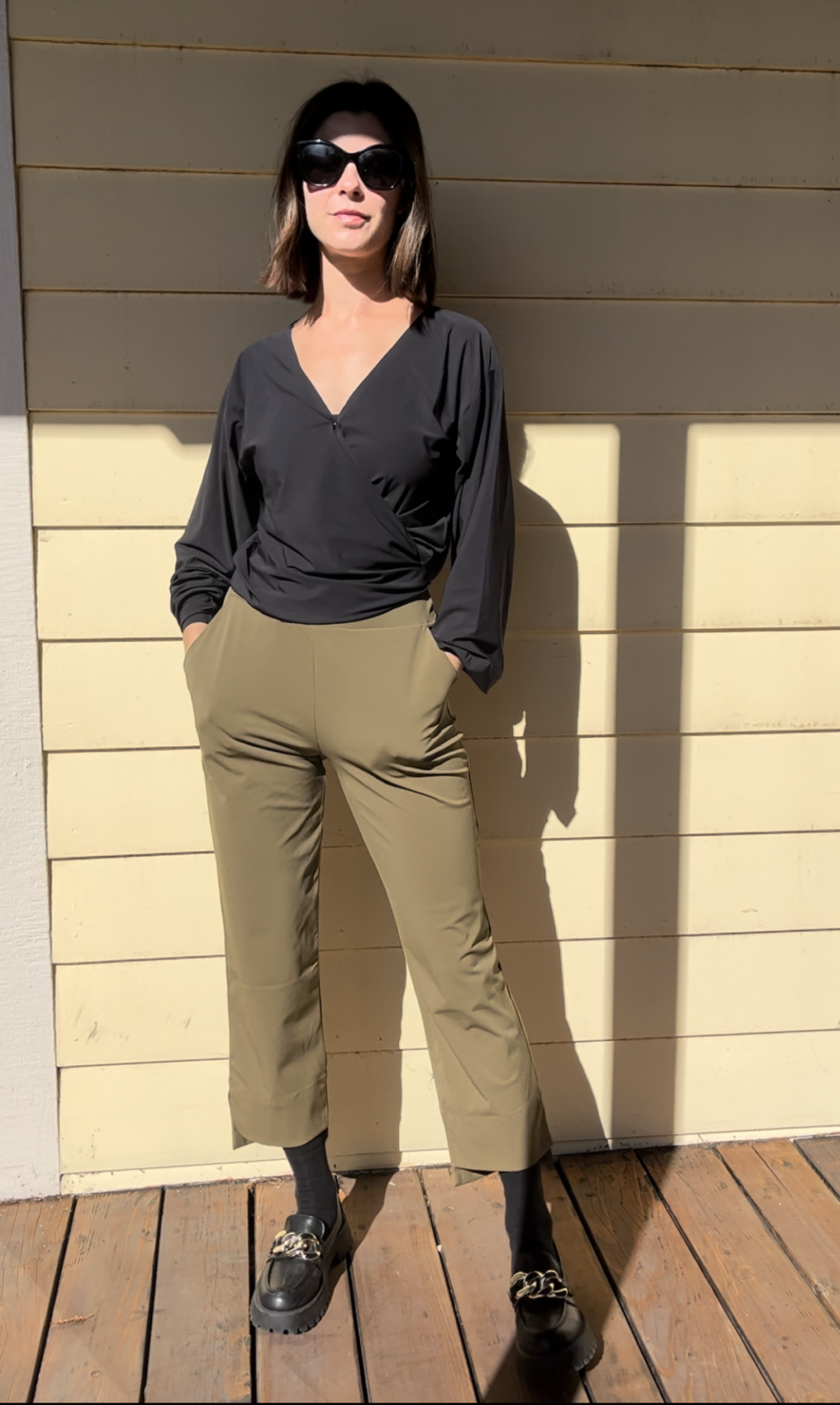Woman standing against a light yellow wooden wall, wearing sunglasses, a black long-sleeve top, olive green pants, and black shoes with gold accents. Sunlight casts shadows behind her.