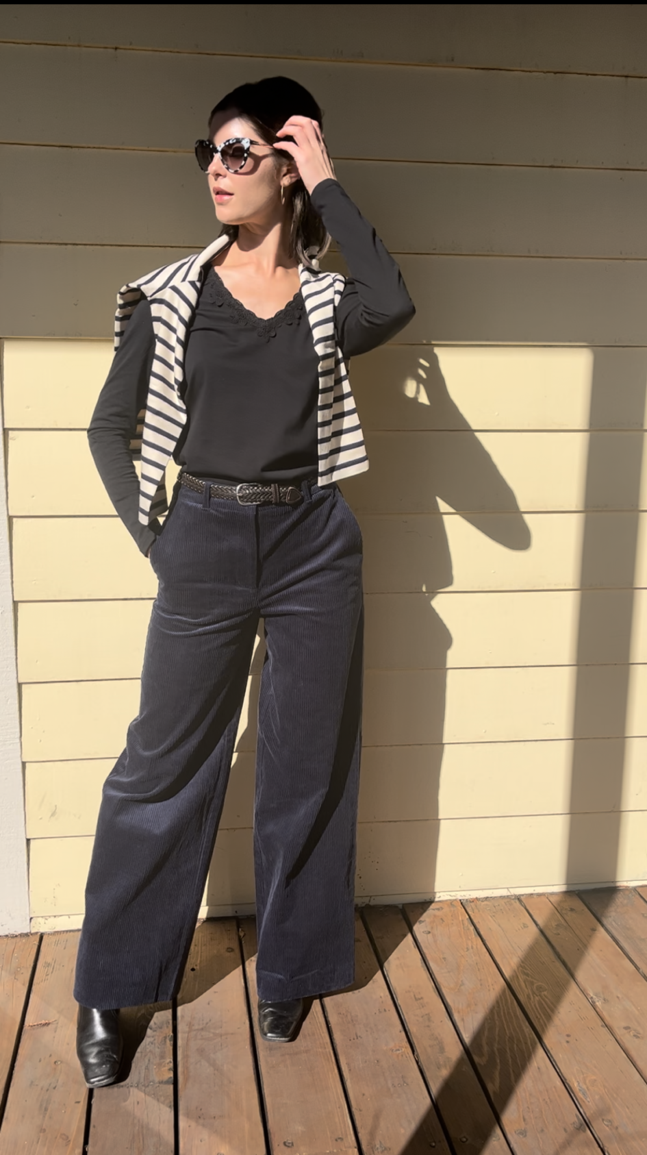 Woman standing on a wooden deck against a light yellow wall, wearing sunglasses, a beret, a striped vest, a black top, wide-leg pants, and black boots.