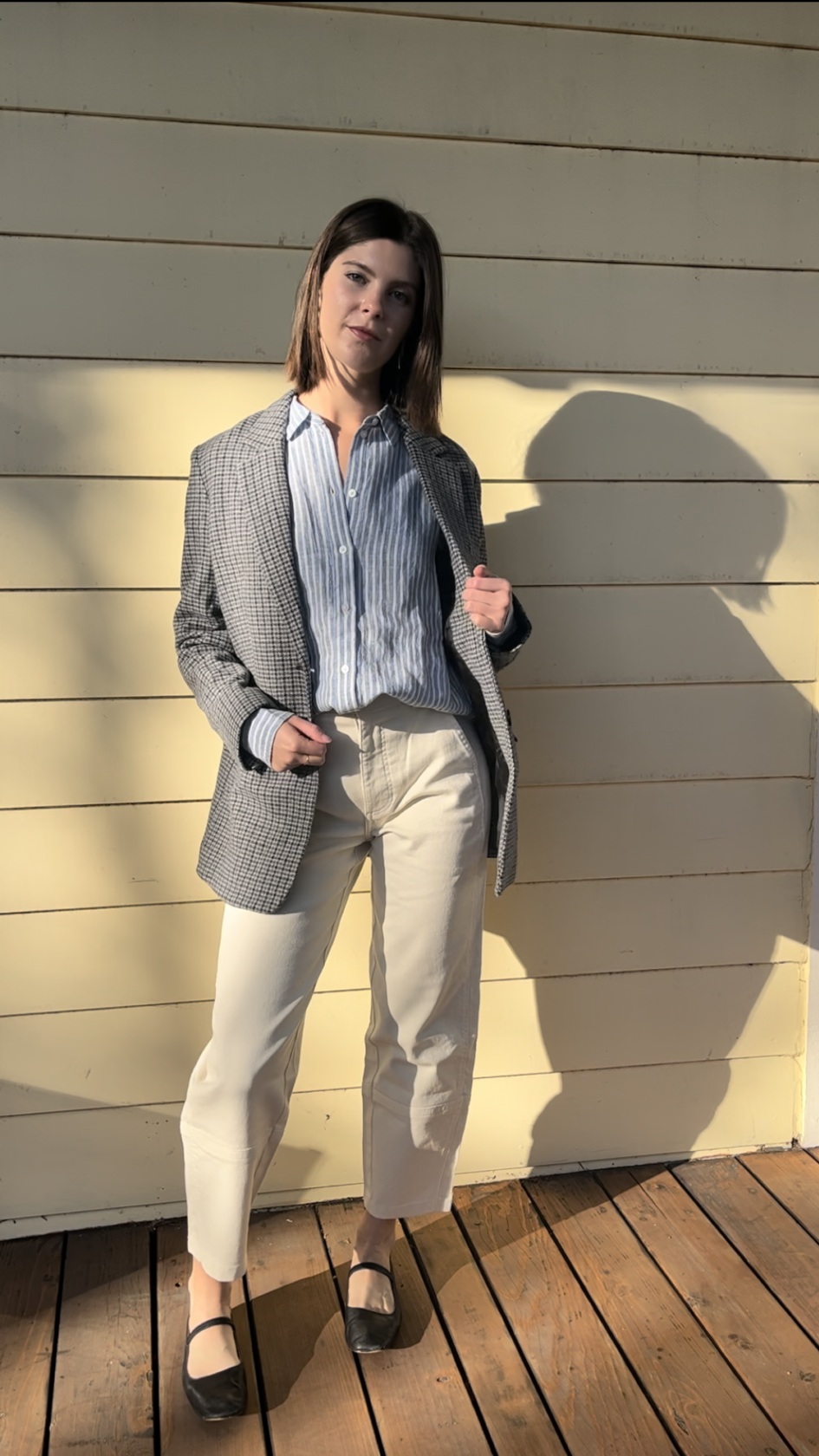 A woman stands on a wooden deck in sunlight, wearing a striped shirt, checkered blazer, light-colored pants, and black shoes, with one hand in her pocket.