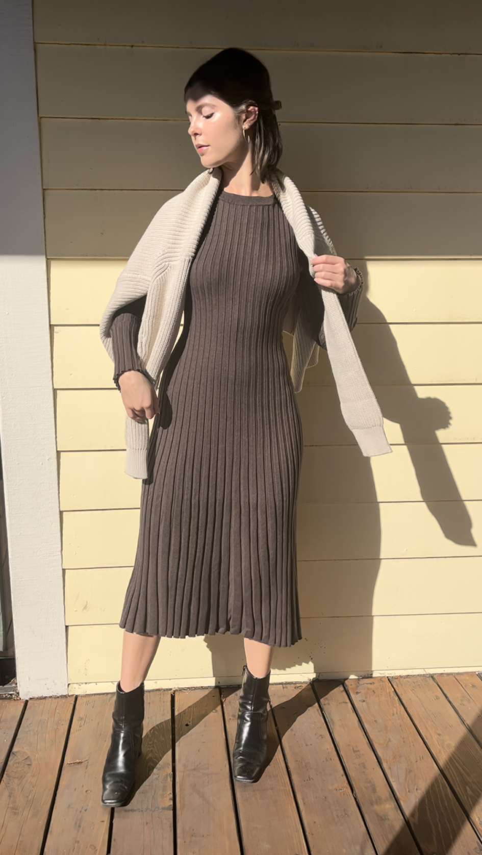 A woman stands on a wooden deck against a yellow wall, wearing a ribbed brown dress, black ankle boots, and a cream sweater draped over her shoulders.