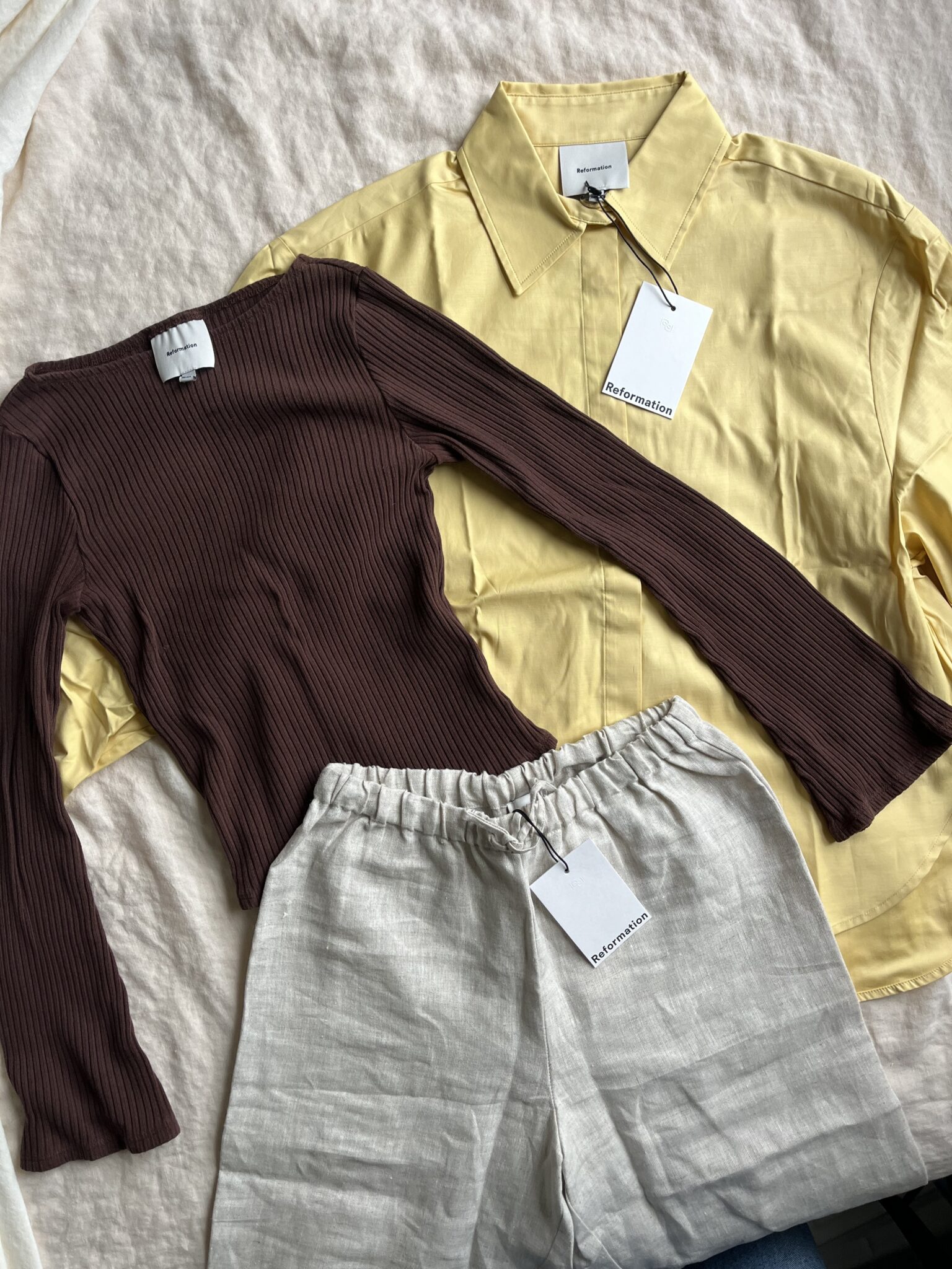 A yellow collared shirt, a brown ribbed long-sleeve top, and beige elastic-waist pants are laid out on a white surface, each with a clothing tag attached.