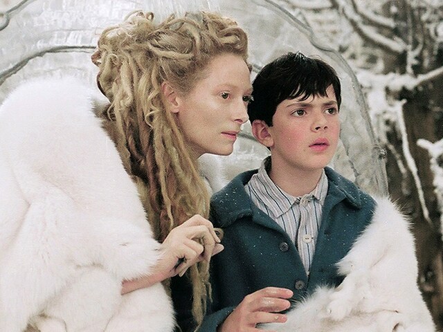A woman with pale skin and long, blond dreadlocks wearing a white fur cloak stands beside a young boy in a blue coat in a snowy outdoor setting.