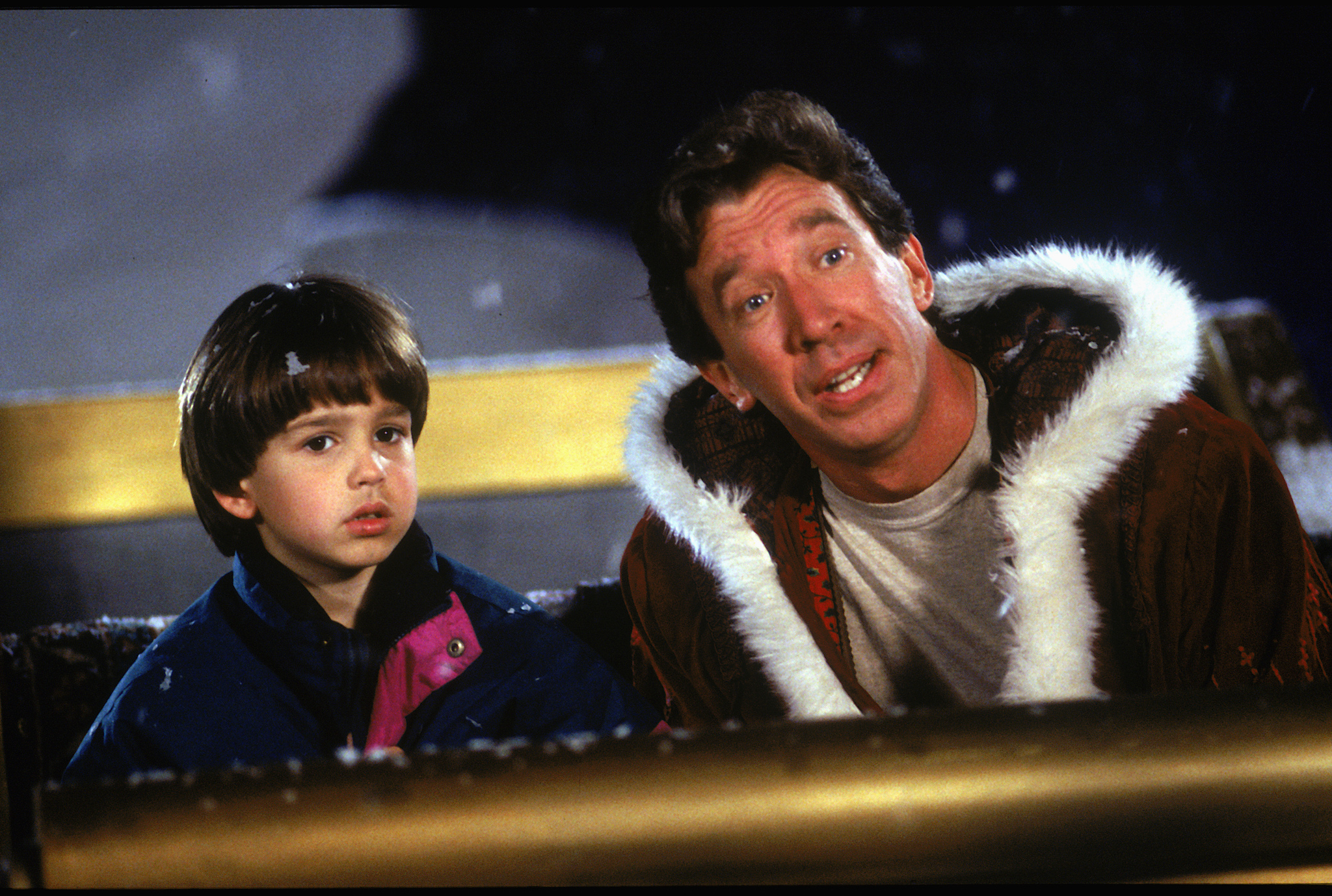 An adult man in a fur-trimmed coat sits next to a young boy in a jacket, both looking ahead while sitting outdoors in a snowy setting.