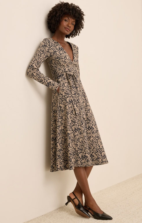 A woman stands against a light wall wearing a long-sleeved, patterned midi dress with a V-neckline and black low-heeled shoes from sustainable brands.