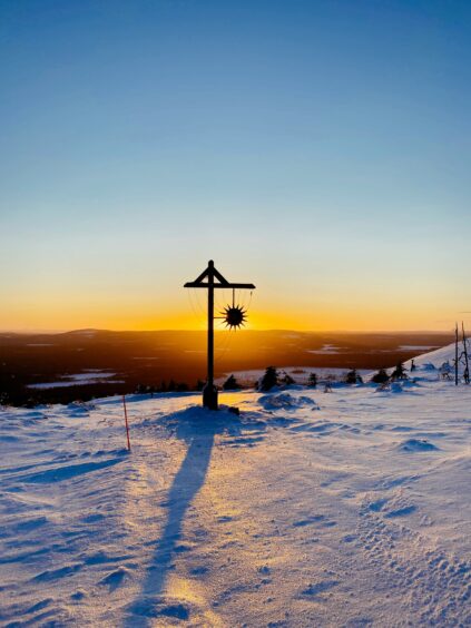 A wooden post with a metal sun ornament casts a long shadow on a snowy landscape at sunset, with distant trees and a clear sky in the background.