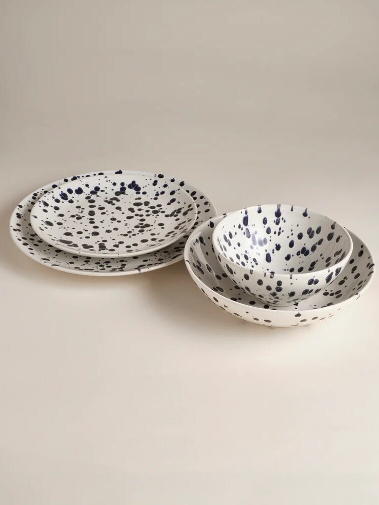 Four ceramic dishes, including two plates and two bowls, with a white base and black splatter pattern, arranged on a neutral background.
