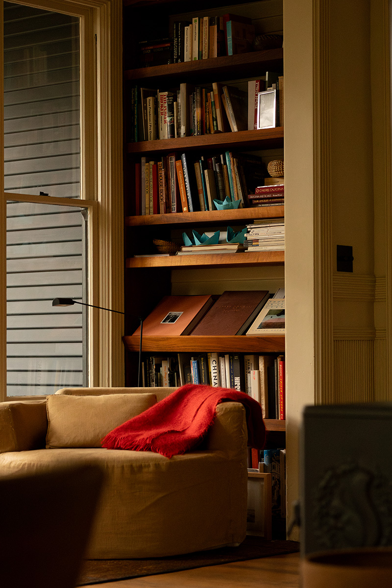 A beige armchair with a red blanket draped over it sits in front of a tall wooden bookshelf filled with books and objects, next to a window.