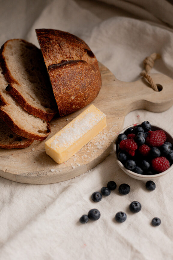 A round loaf of sliced bread, a block of cheese, and a small bowl of blueberries and raspberries on a wooden board, with a few berries scattered on a beige cloth.