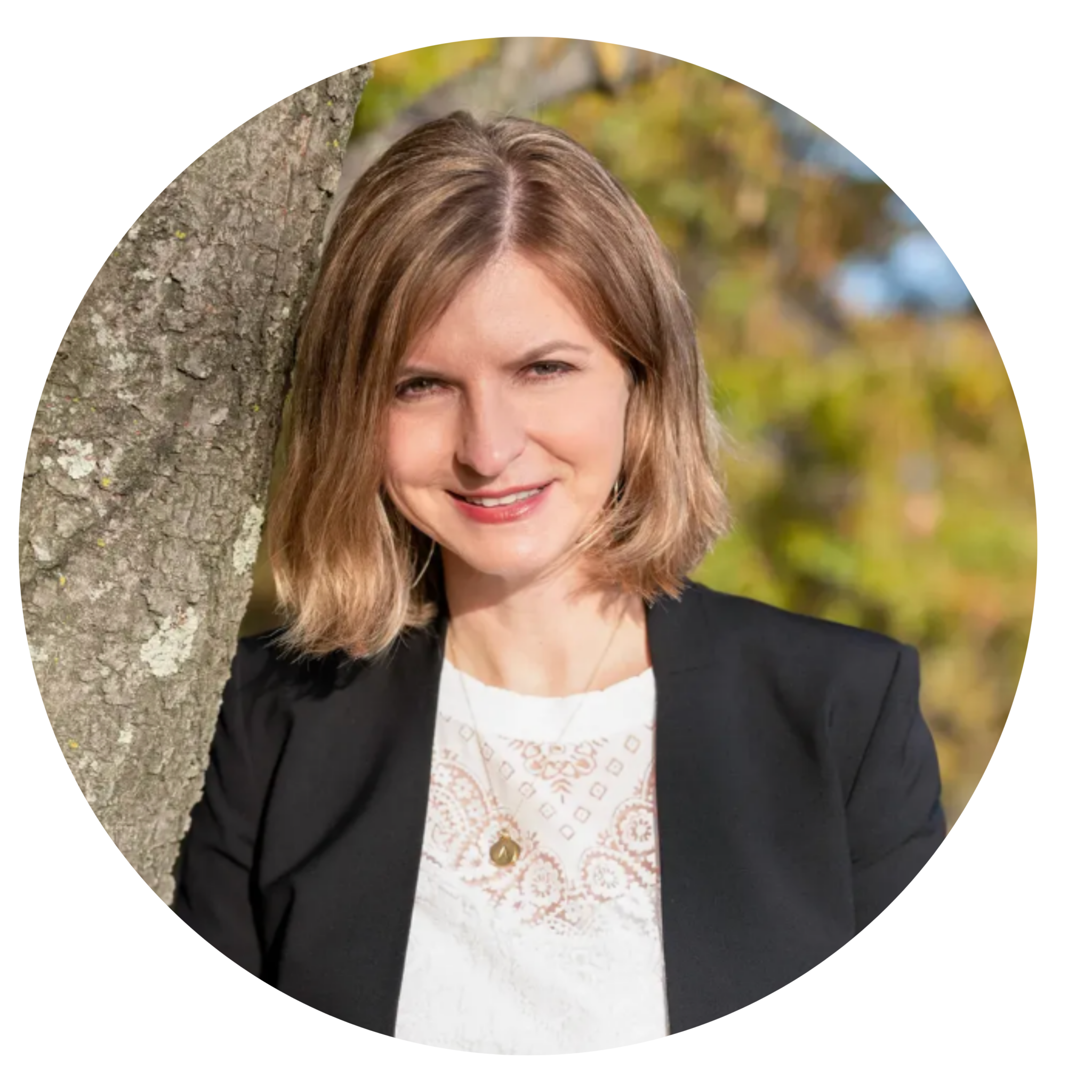 A woman with shoulder-length blonde hair, wearing a black blazer and white top, stands outdoors next to a tree with blurred greenery in the background.
