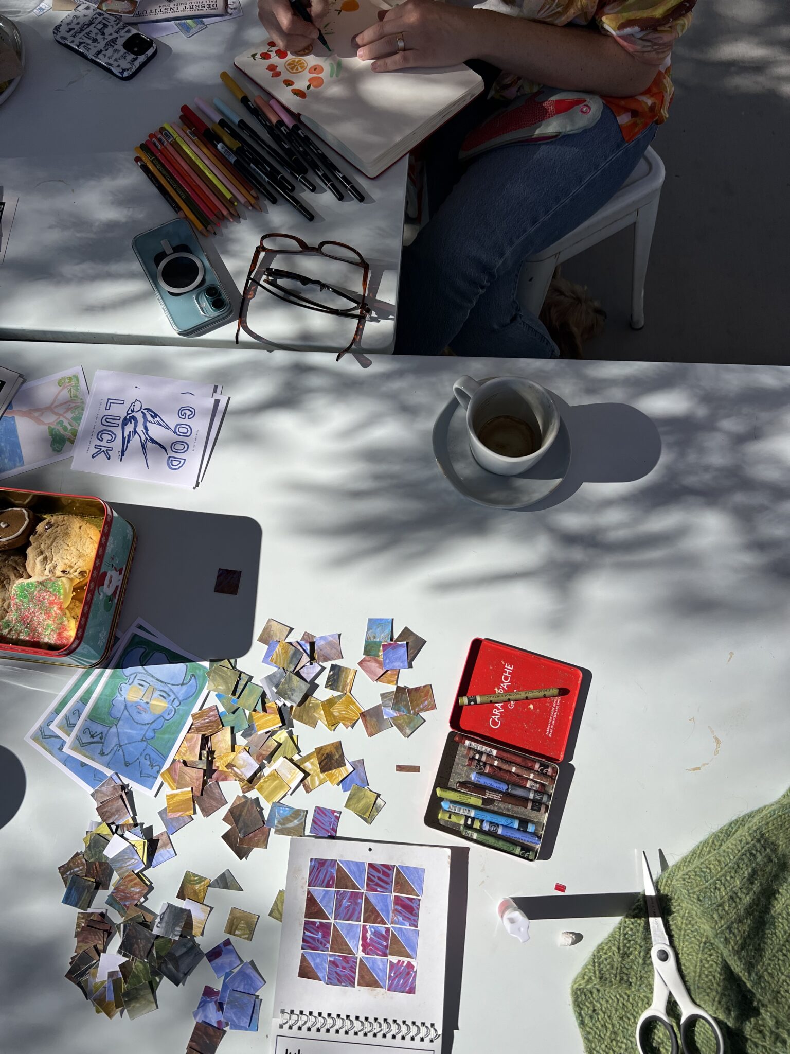 A person arranges small painted paper squares on a white table with art supplies, a coffee cup, scissors, and sunlight casting shadows across the scene.