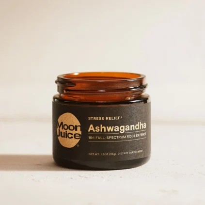 A brown glass jar labeled "Moon Juice Ashwagandha 15:1 Full-Spectrum Root Extract" sits on a light surface. The label notes it is a dietary supplement for stress relief.