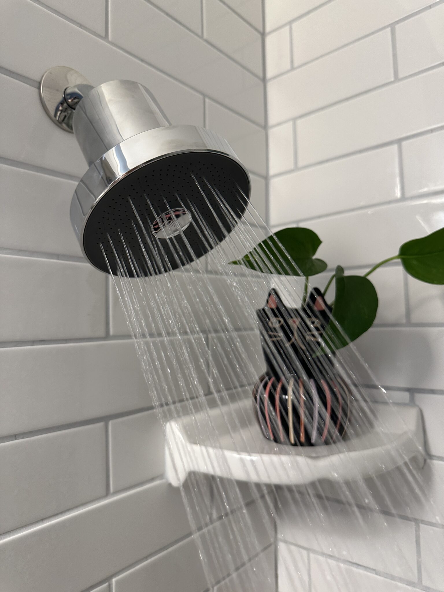 A chrome showerhead sprays water onto a white corner shelf holding a decorative vase with a green plant, set against white subway tile walls.