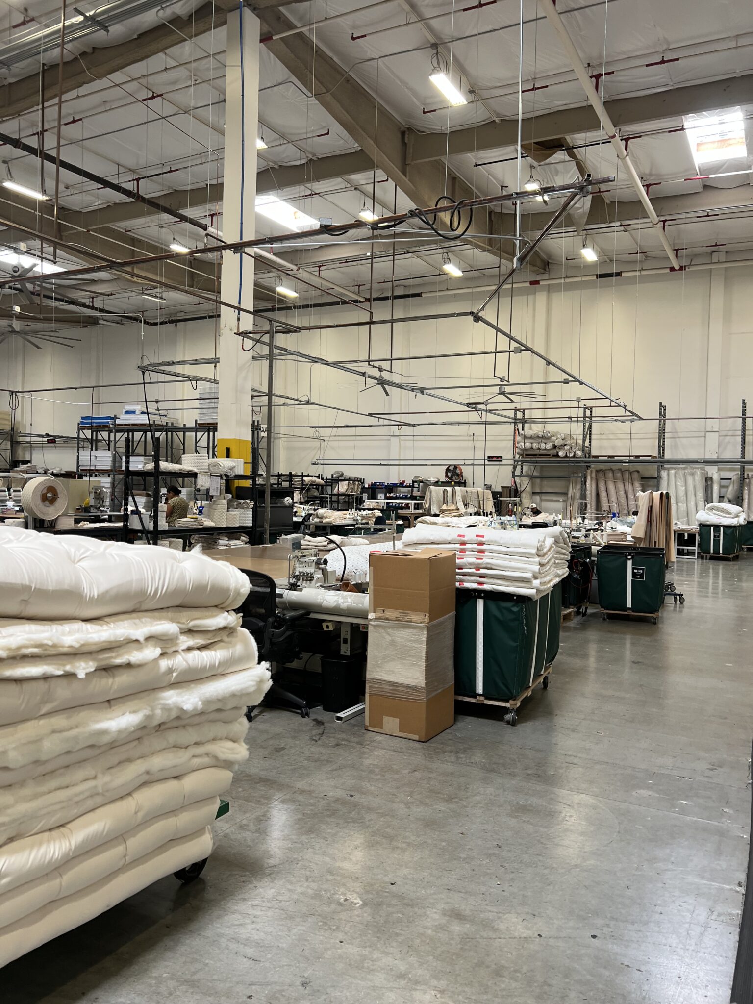 A textile factory floor with stacked fabric rolls, industrial sewing machines, workstations, and carts, under bright overhead lighting.