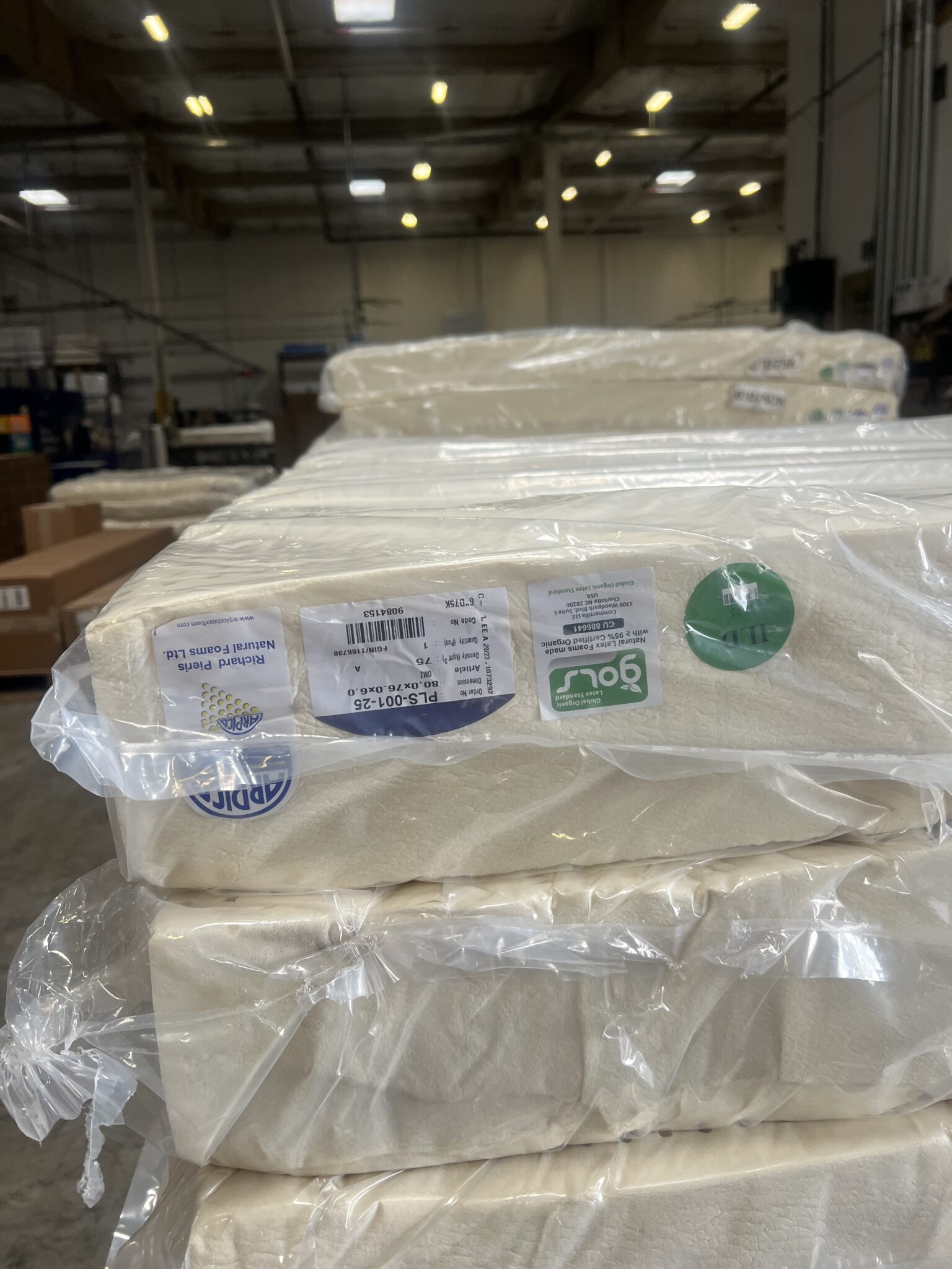 Stacks of foam cushions wrapped in plastic, labeled with product and recycling information, are placed inside a warehouse with high ceilings and industrial lighting.