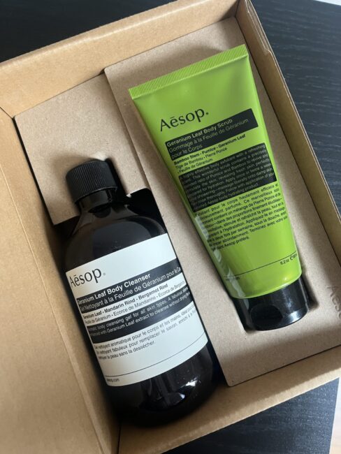 Aesop body cleanser and green tube of hand balm in a brown cardboard box on a black surface.