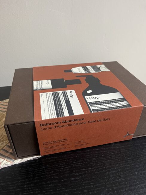A brown Aesop gift box labeled "Bathroom Abundance" sits on a dark wooden surface next to a beige cloth.