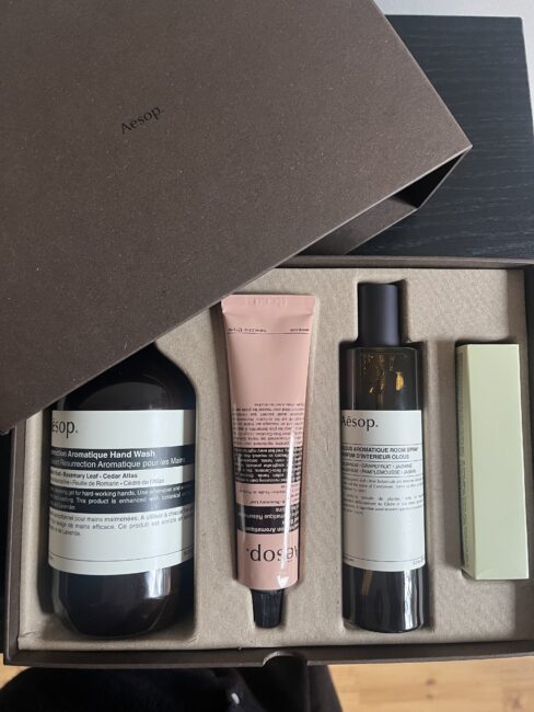 A brown Aesop gift box containing an aromatic hand wash, a pink tube of hand balm, a facial mist, and a rectangular box, all neatly arranged in molded packaging.