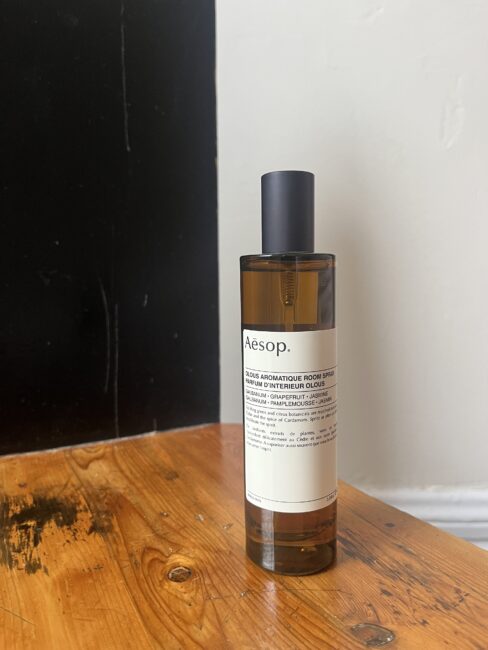 A brown bottle of Aesop room spray sits on a wooden surface near a black and white wall.