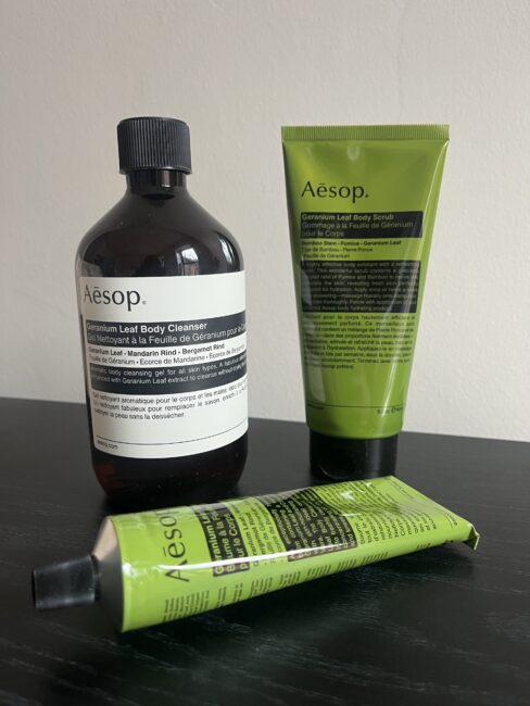 Three Aesop Geranium Leaf body care products, including a bottle of body cleanser, a tube of body scrub, and a tube of body balm, displayed on a dark surface against a light background.