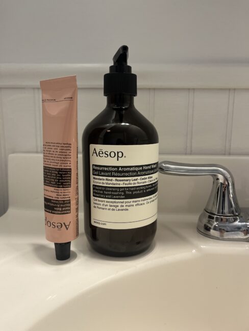 A tube of Aesop hand cream and a bottle of Aesop Resurrection Aromatique Hand Wash are placed on a bathroom sink next to a silver faucet.