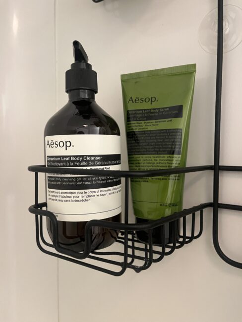 A brown pump bottle and a green tube of Aesop Geranium Leaf products sit in a black metal shower caddy against a white tiled wall.