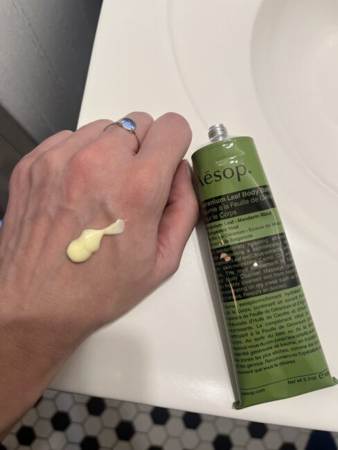 A hand with a dab of yellowish lotion on the back is next to a green tube of Aesop Geranium Leaf Body Balm on a bathroom sink.