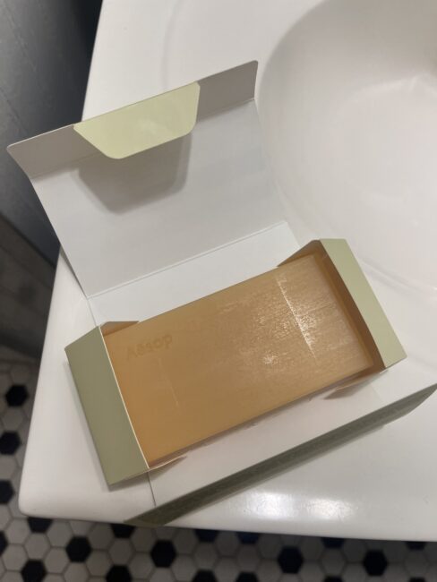 A rectangular bar of Aesop soap in an open cardboard box is placed on a white bathroom counter next to a sink.