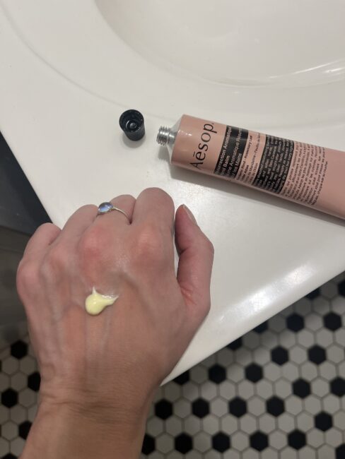 A hand with a small amount of yellow cream on the back, next to an open Aesop tube on a white bathroom sink.