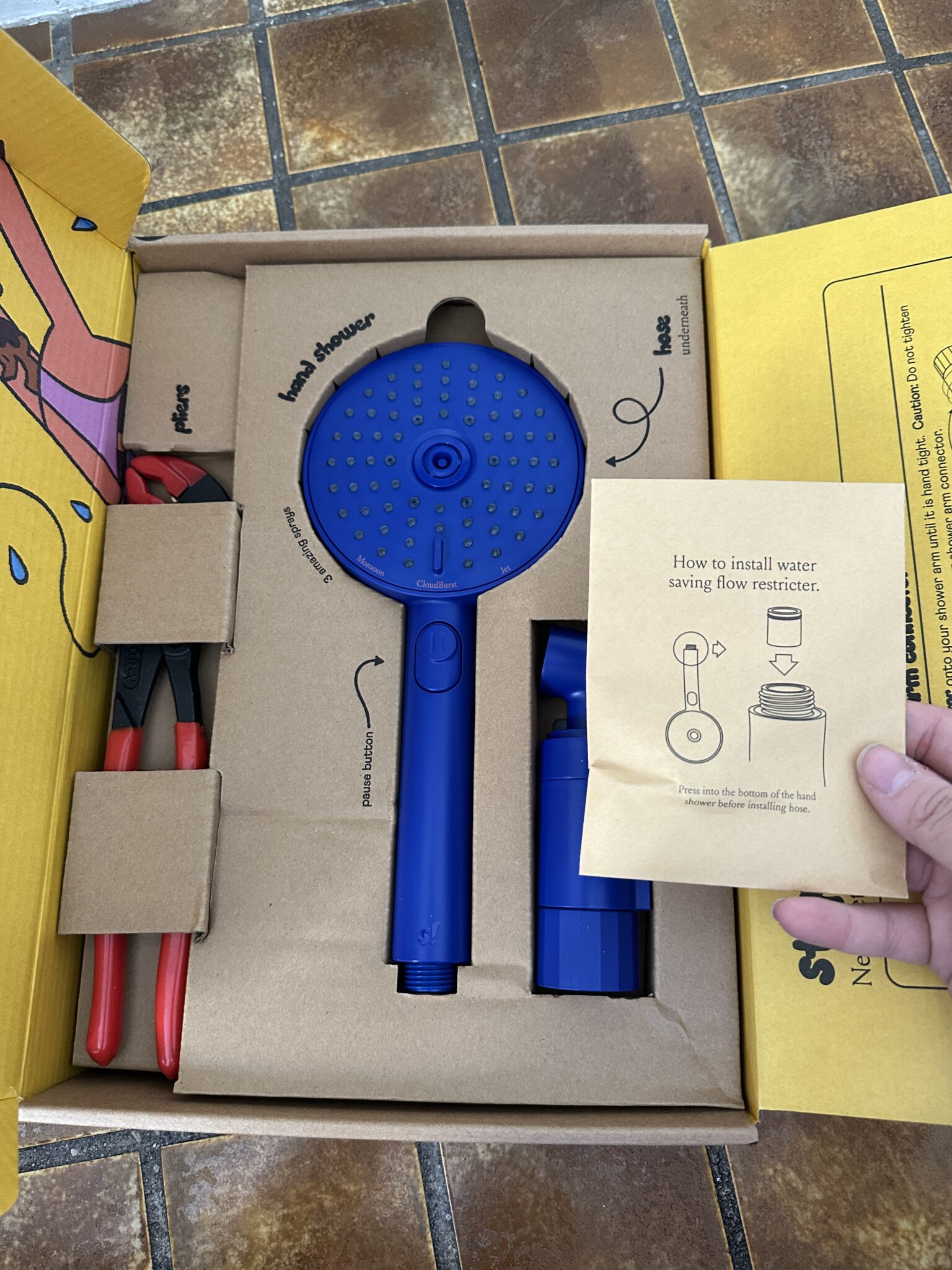 A blue showerhead kit with installation instructions and red-handled pliers inside a yellow box on a tiled surface.
