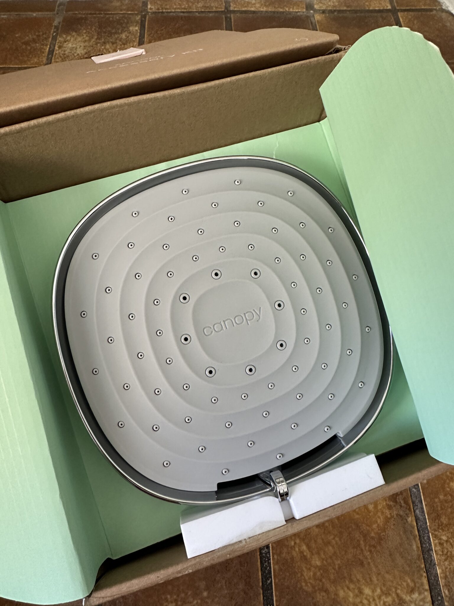 A Canopy brand showerhead in a cardboard box with light green interior flaps, resting on a tiled surface.