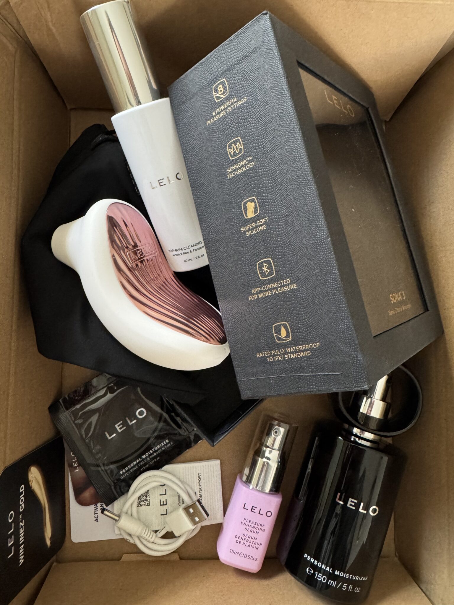 A box containing LELO branded items including a personal massager, toiletry products, a charging cable, sachets, a pouch, and product packaging.