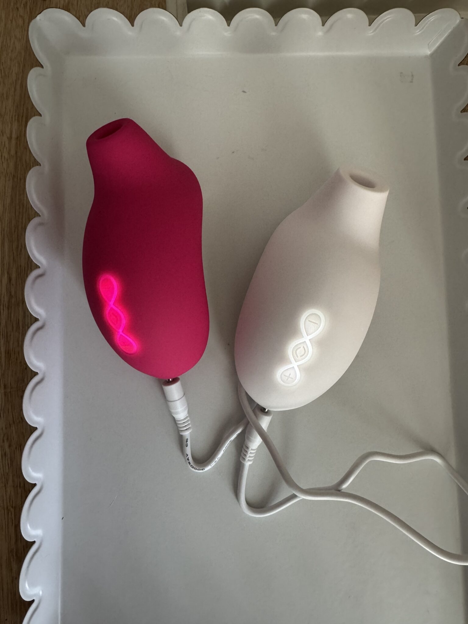 Two silicone suction vibrators, one pink and one white, are placed on a tray and plugged in with charging cables. Both devices have illuminated control symbols.