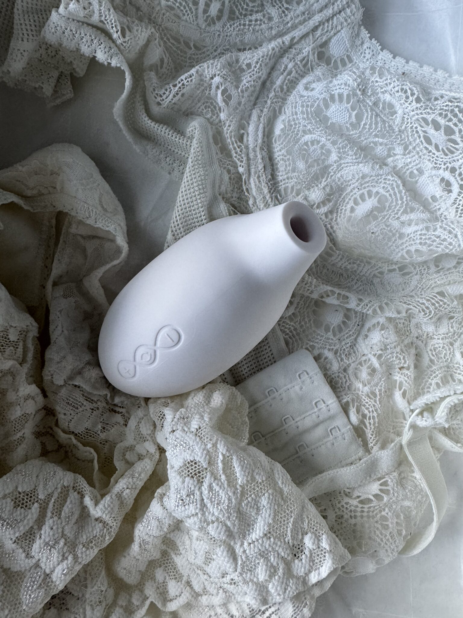 A small, white ceramic vase with simple embossed design resting on white lace fabric and lingerie.