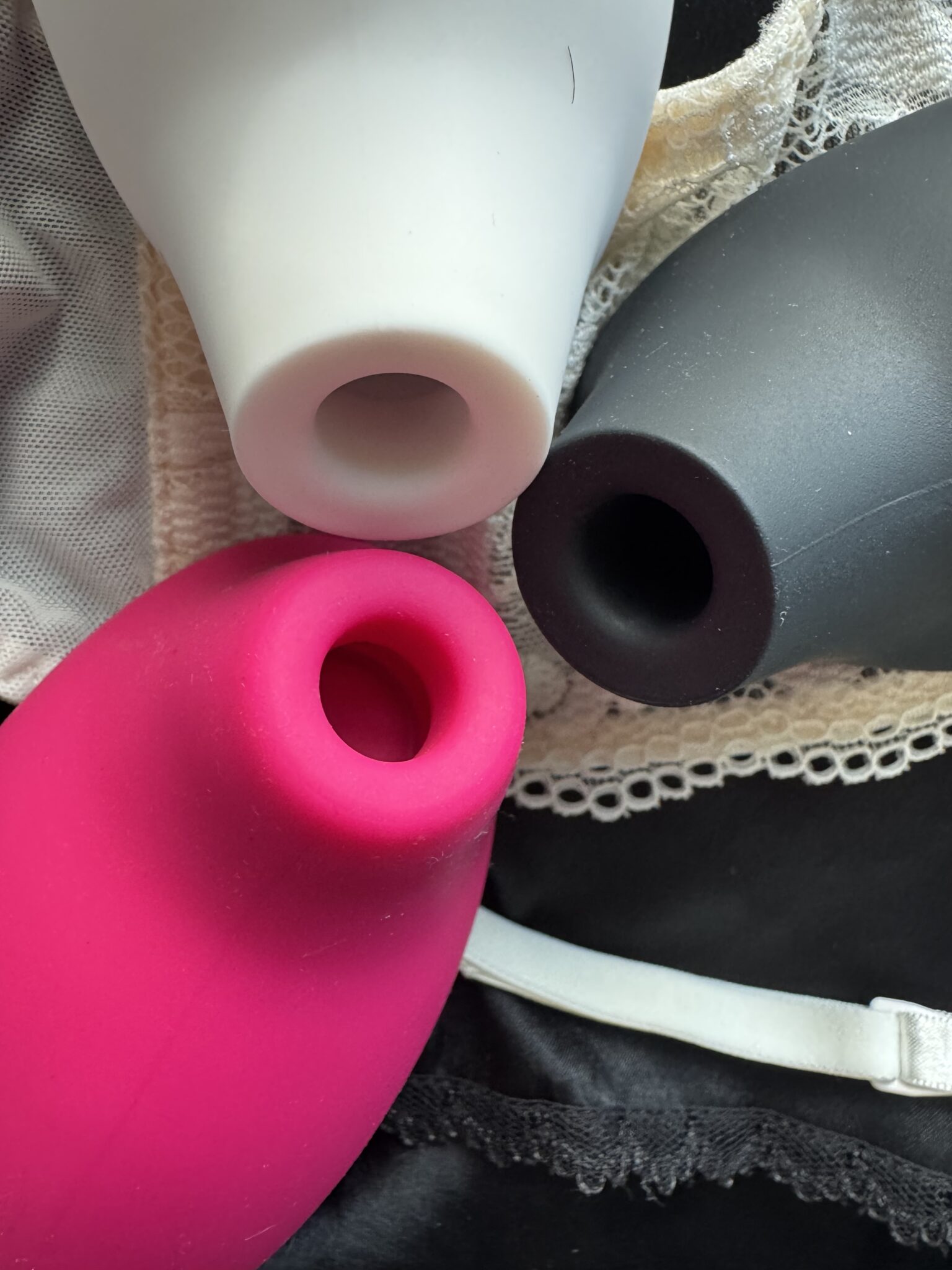 Three silicone suction devices in white, black, and pink are arranged closely together on lace and satin fabric.