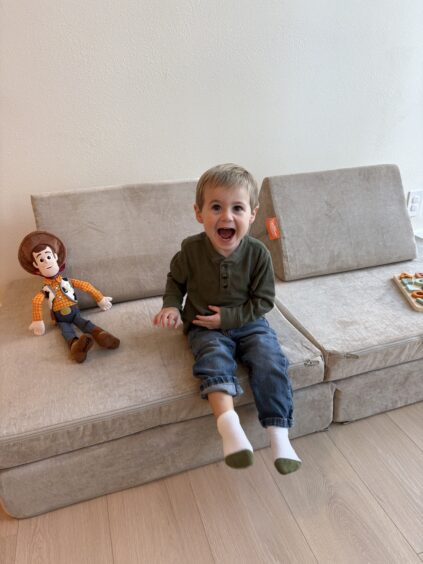 A young boy with light brown hair sits on a beige couch, smiling widely. Next to him is a Woody doll from Toy Story, and a tray with snacks is on the couch.