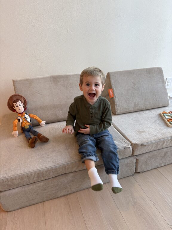 A young boy with light brown hair sits on a beige couch, smiling widely. Next to him is a Woody doll from Toy Story, and a tray with snacks is on the couch.
