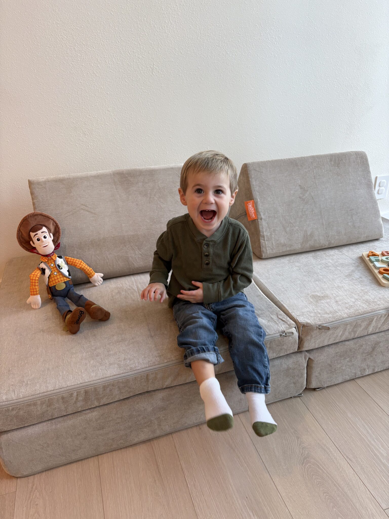 A young boy with light brown hair sits on a beige couch, smiling widely. Next to him is a Woody doll from Toy Story, and a tray with snacks is on the couch.
