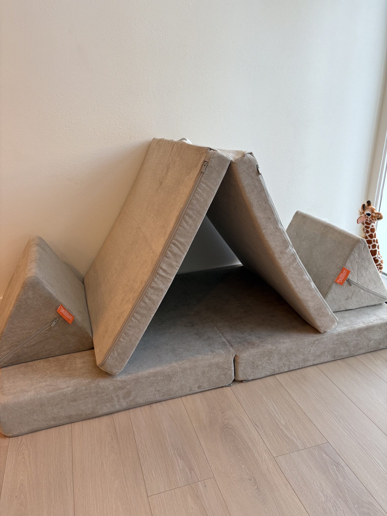 A foldable foam play couch is arranged as a tent with two triangular cushions on the sides. A toy giraffe is visible in the background near the wall.
