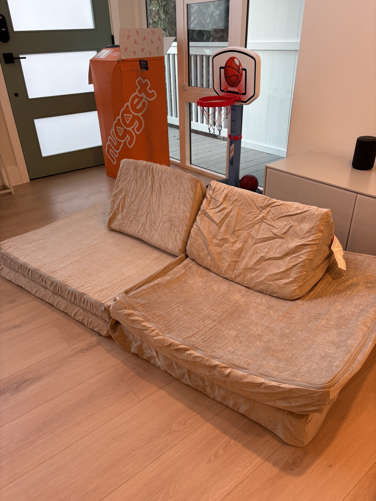 Two tan foam cushions set up as a makeshift couch next to a Nugget box, a small basketball hoop, and a white cabinet in a living room.