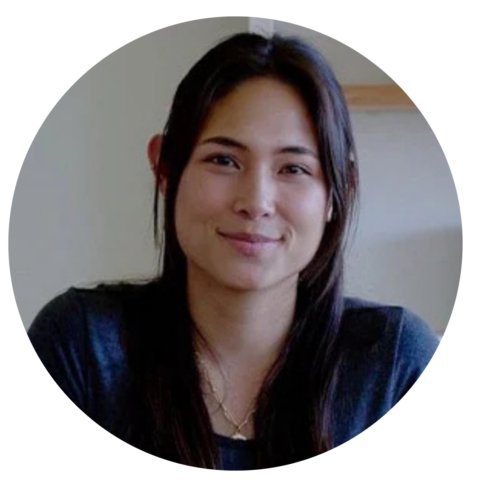 A person with long dark hair and a dark top, sitting indoors and looking at the camera with a slight smile.