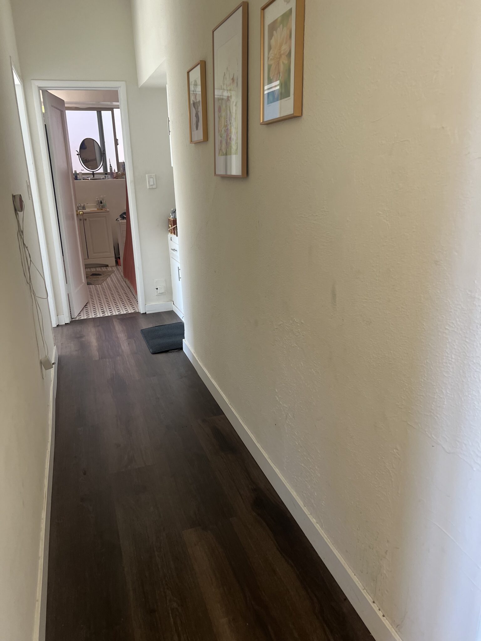 A narrow hallway with dark wood flooring, white walls, framed pictures, and an open door at the end leading to a room with a sink and mirror.