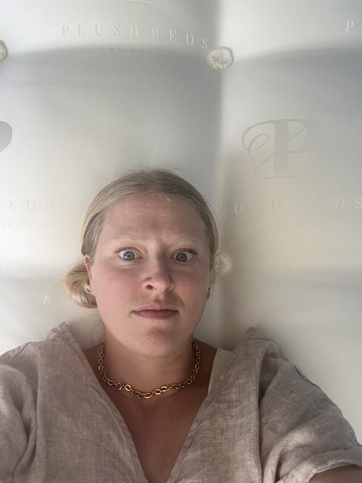 A woman with blonde hair and a neutral expression lies on a mattress with "PlushBeds" branding visible in the background.