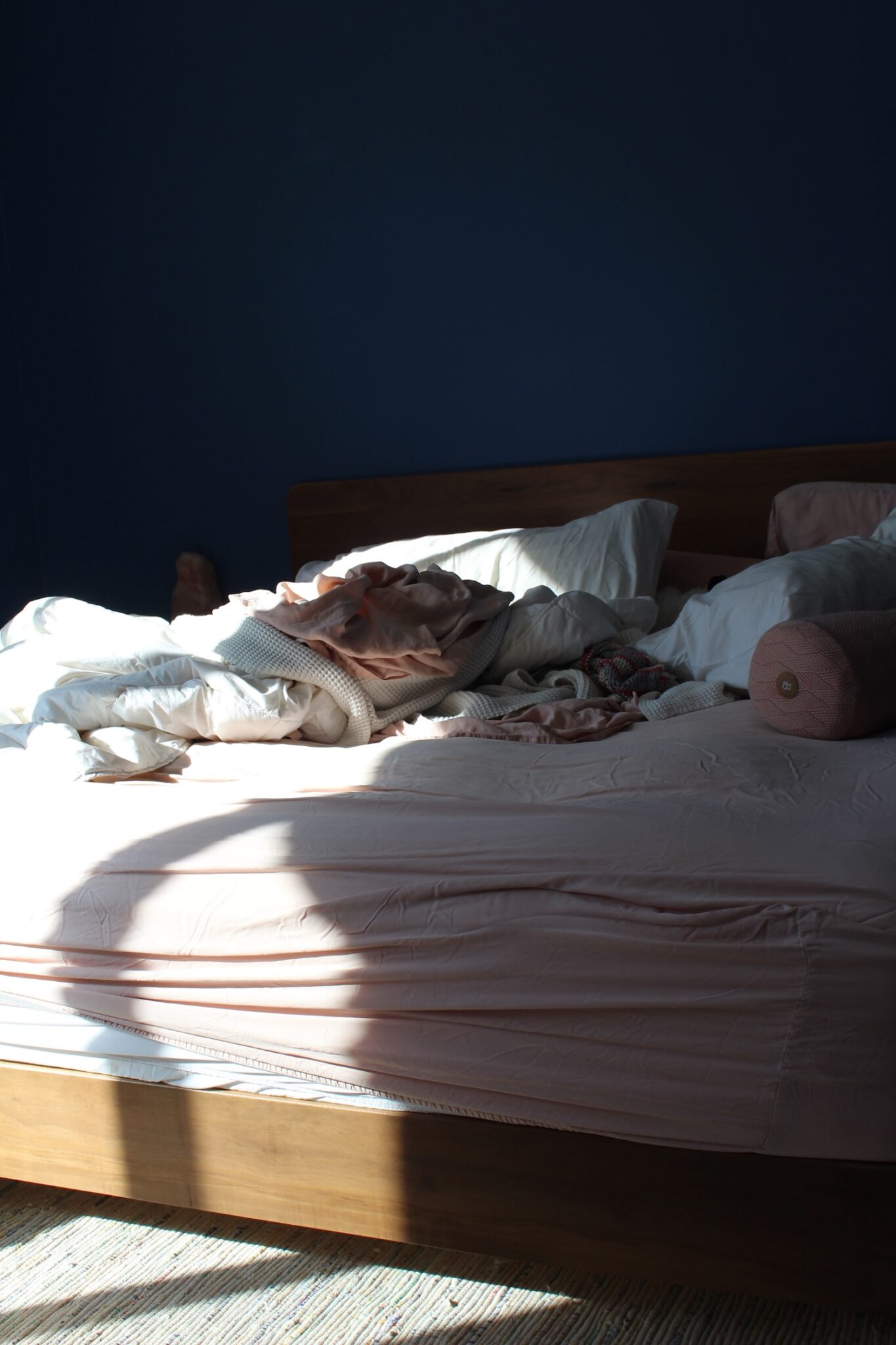A messy bed with rumpled sheets, blankets, and pillows is bathed in natural sunlight against a dark blue wall.