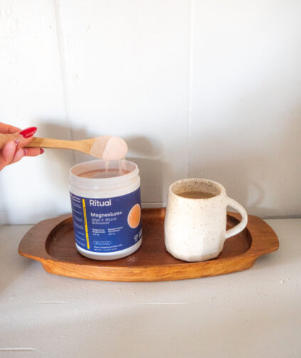 A hand scoops powder from a Ritual Magnesium+ container next to a ceramic mug on a wooden tray.