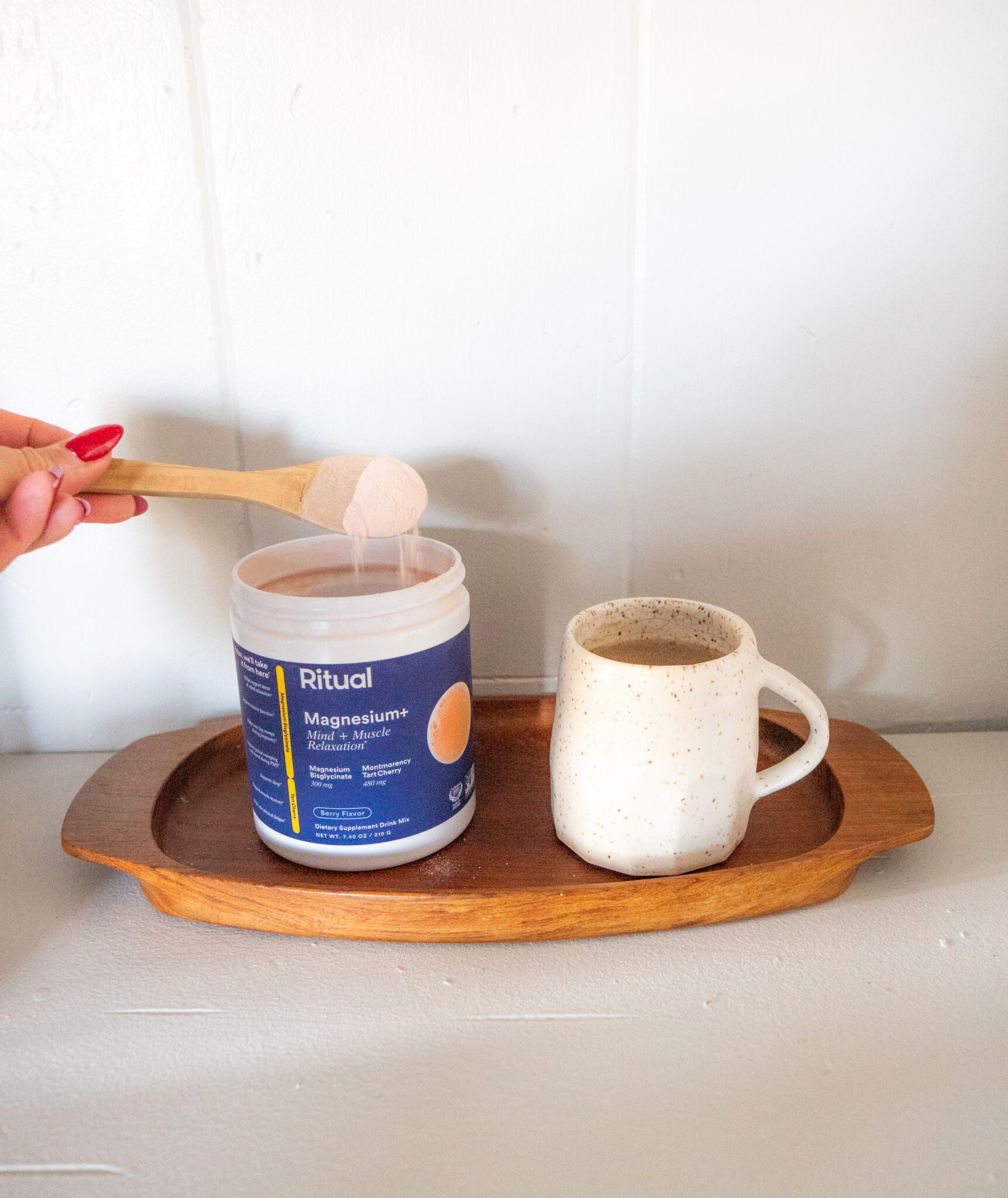 A hand scoops powder from a Ritual Magnesium+ container next to a ceramic mug on a wooden tray.