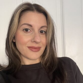 A woman with straight, shoulder-length brown hair with blonde highlights, wearing a black top, poses in front of a white paneled wall.