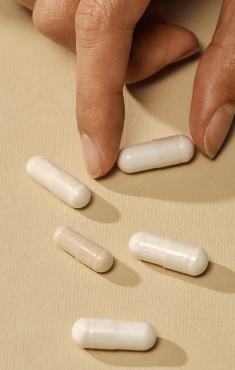 A hand reaching toward white capsules scattered on a beige surface.