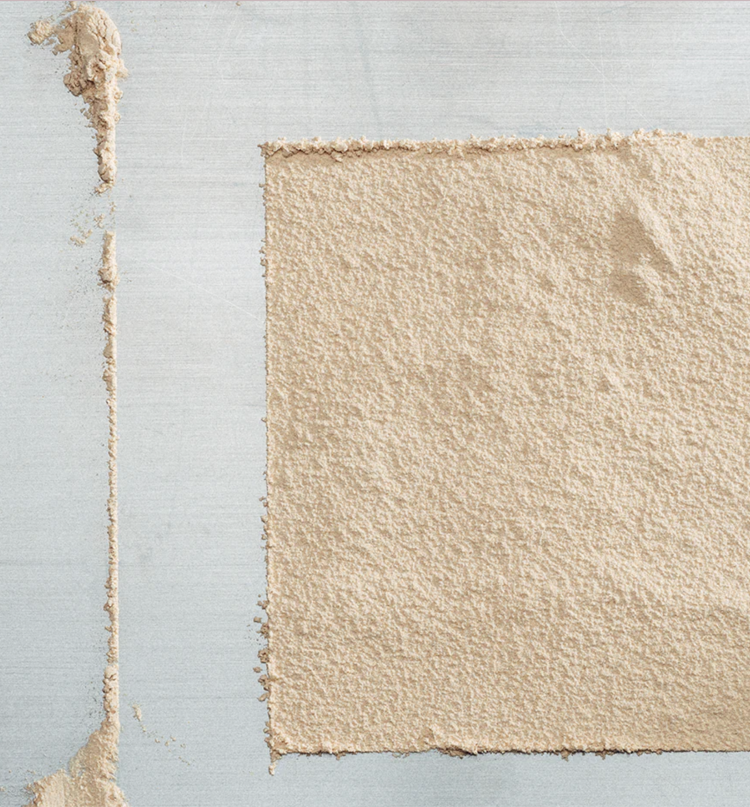 A square area of light beige powder spread evenly on a flat, light-colored surface, with excess powder along the edges.