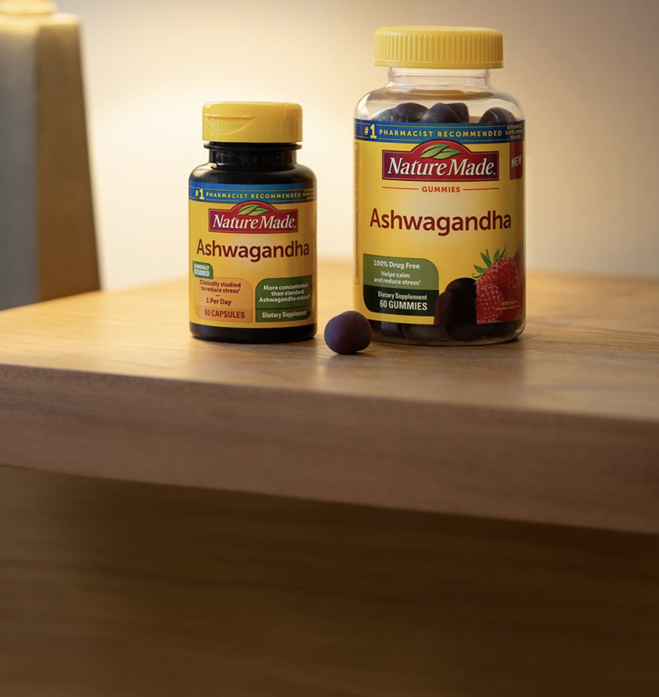 Two bottles of Nature Made Ashwagandha supplements, one with capsules and one with gummies, are placed on a wooden surface with a soft light background.